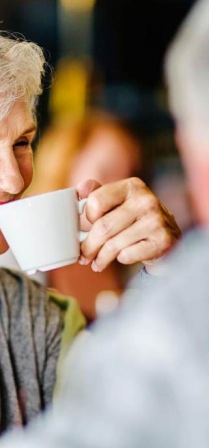 Elderly woman smiling and holding a cup, sitting across from a man.