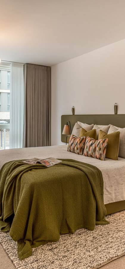 A modern bedroom with a large bed, green and white decor, a desk, chairs, and a window opening to a balcony.