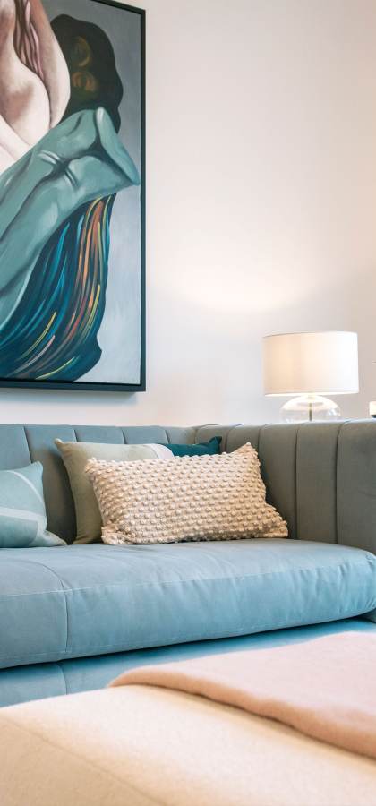 A modern living room featuring a teal sofa with cushions and a beige throw, a wall with abstract artwork, a lit table lamp, and a coffee table with decor items including a pink vase and reed diffuser.