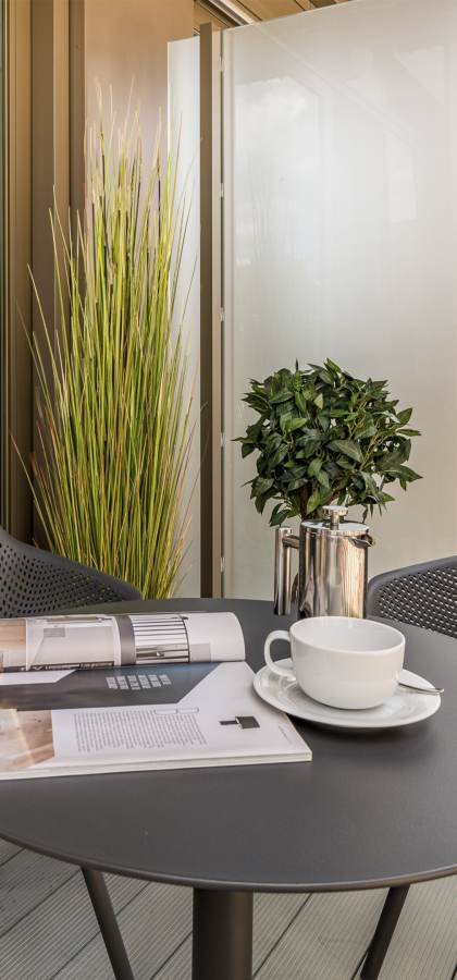 A small outdoor balcony with two black chairs and a round table. On the table are a white cup and saucer, two books, and a plant in a silver pot. The setup overlooks a building.