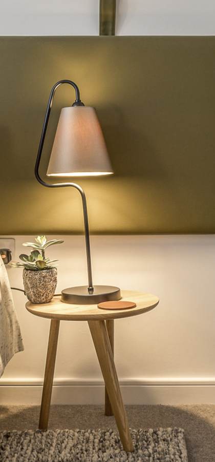 A modern bedroom features a neatly made bed, a bedside table with a lamp and a small plant, and a brown upholstered chair. The room has a minimalist design with earthy tones.
