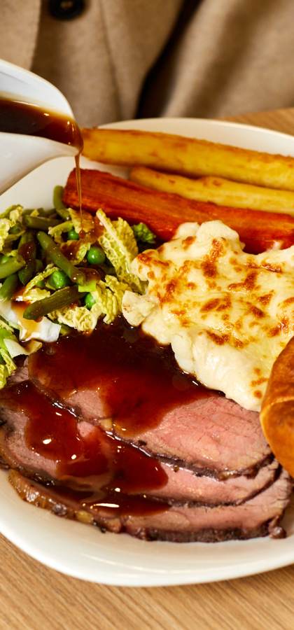 A person pours gravy onto a plate with roast beef, Yorkshire pudding, roast potatoes, carrots, cabbage, and cauliflower cheese. A fork and knife are on a napkin beside the plate.