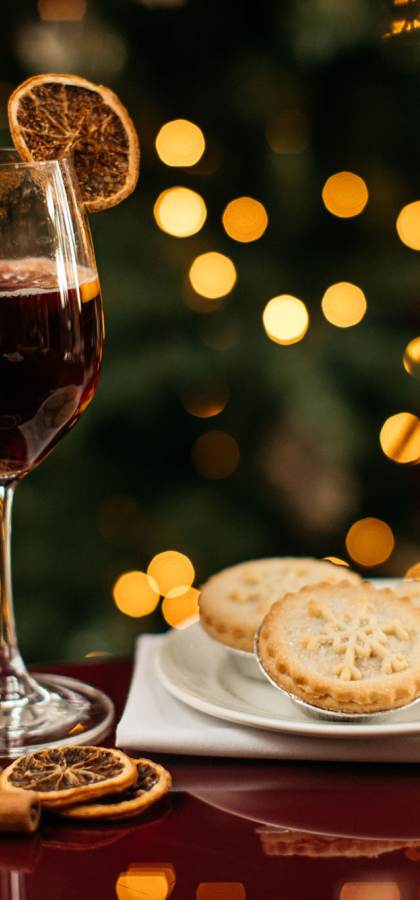 A glass of red wine with a dried orange slice garnish, a small plate of mince pies, cinnamon sticks, and dried orange slices on a red table with blurred lights in the background.