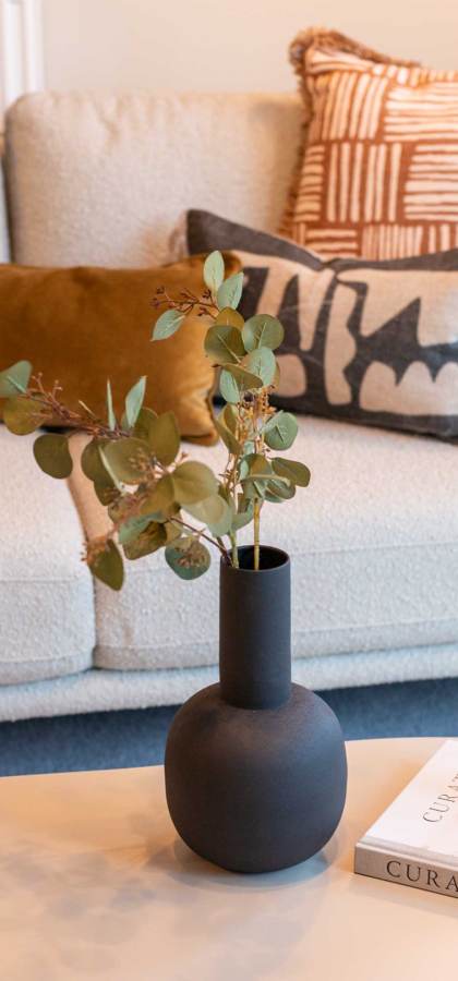 A beige sofa with decorative cushions, and a coffee table with a candle, plant in a black vase, and a reed diffuser atop two books.