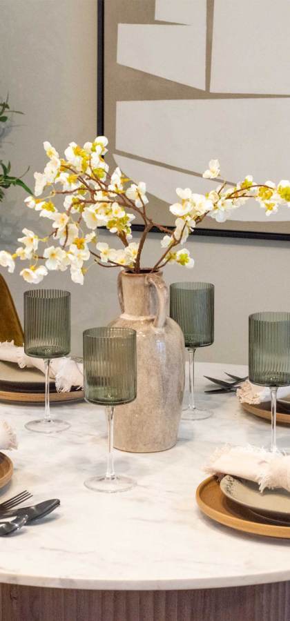 A round dining table set for four with rustic plates, green glasses, and napkins. A vase with white flowers is in the center. Greenery and abstract art decorate the background.