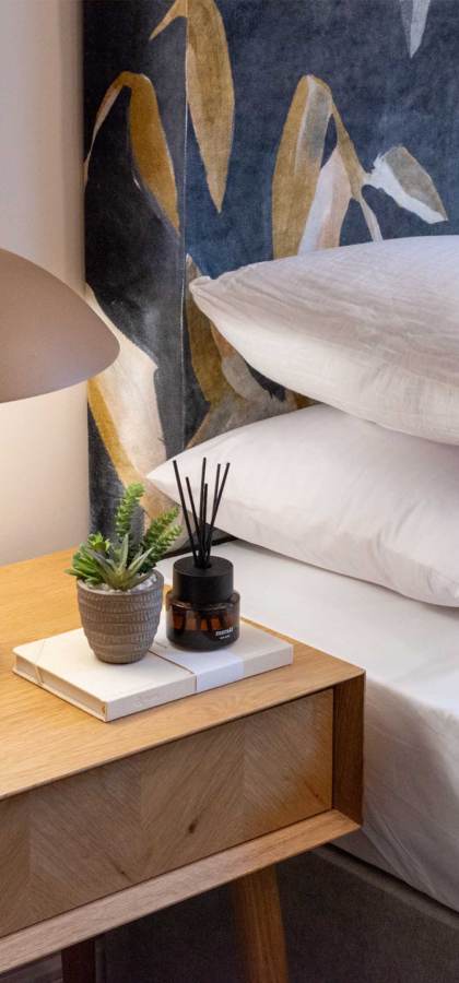 Modern bedroom scene with a wooden nightstand holding a brown lamp, two books, a small plant, and a reed diffuser. The bed has white sheets, multiple pillows, and a decorative wall tapestry.
