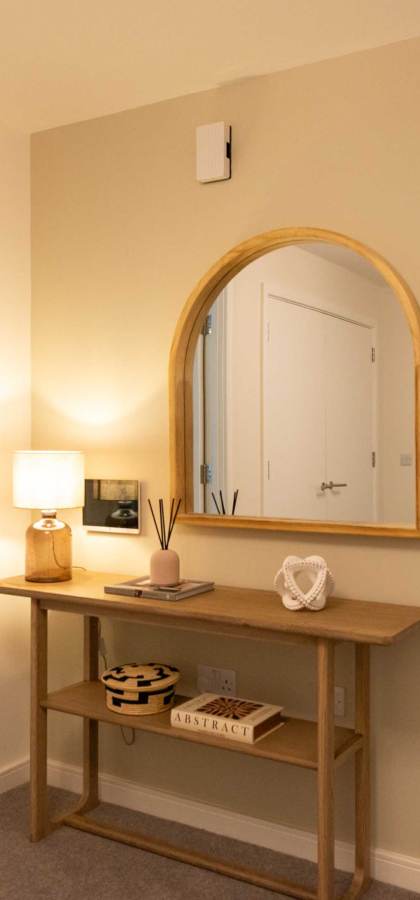A hallway with a wooden console table and mirror. To the left is a dining area with a set table, and to the right is a bedroom with a made bed and large window.