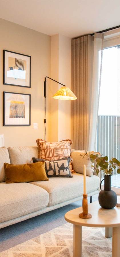 Modern living room with a beige sofa, patterned cushions, and a wooden coffee table. Art prints hang on the wall. A lamp illuminates the space. Balcony with seating is visible through glass doors.