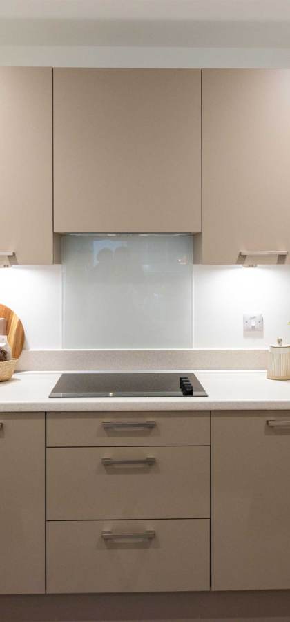 Modern kitchen with beige cabinets, built-in oven, electric cooktop, and white countertops. Plants and kitchenware are placed on the countertops and shelves.