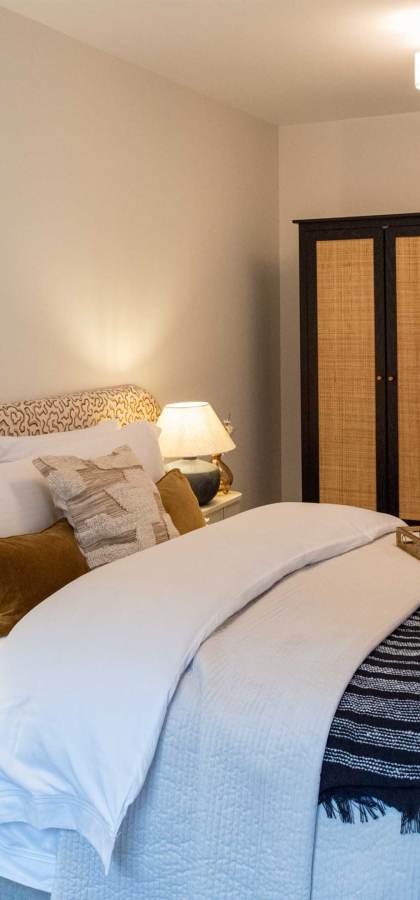 Modern bedroom with a neatly made double bed, patterned headboard, bedside tables with lamps, a dark wardrobe, and a ceiling light. A folded throw is placed at the foot of the bed.
