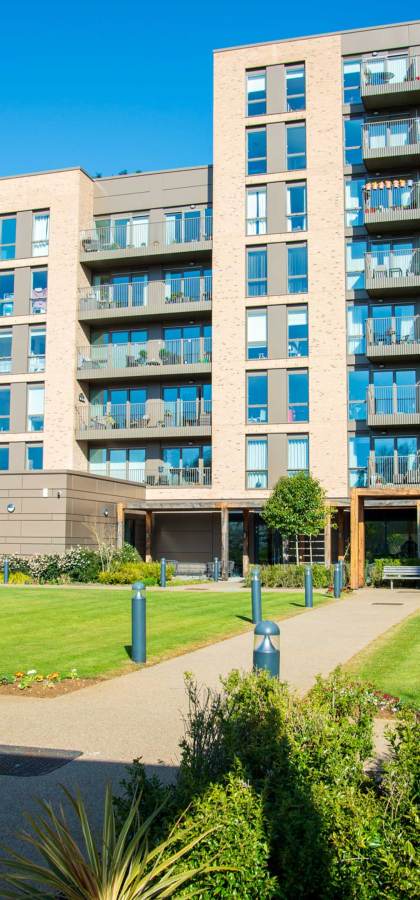 Modern apartment building with several floors, balconies, and large windows, surrounded by a well-maintained garden and pathways under a clear blue sky.