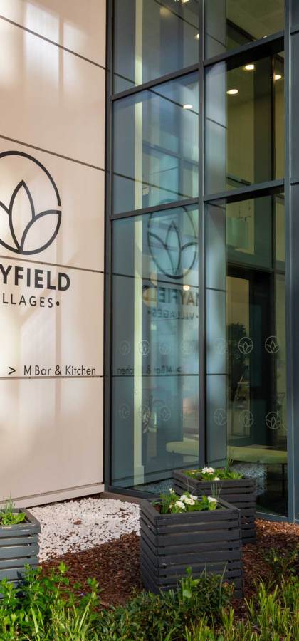 Exterior of Mayfield Villages building entrance with glass facade, plants in planters, and visible sign for M Bar & Kitchen.