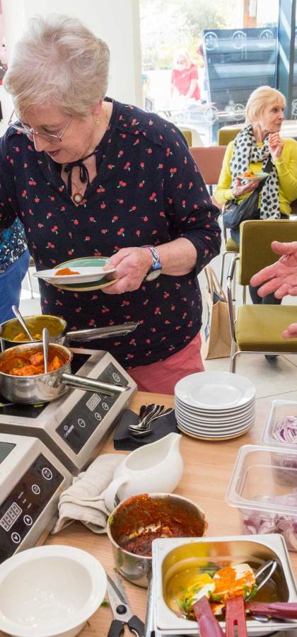 People serving themselves food from a buffet table with various dishes, bowls, and cooking equipment in a casual setting.