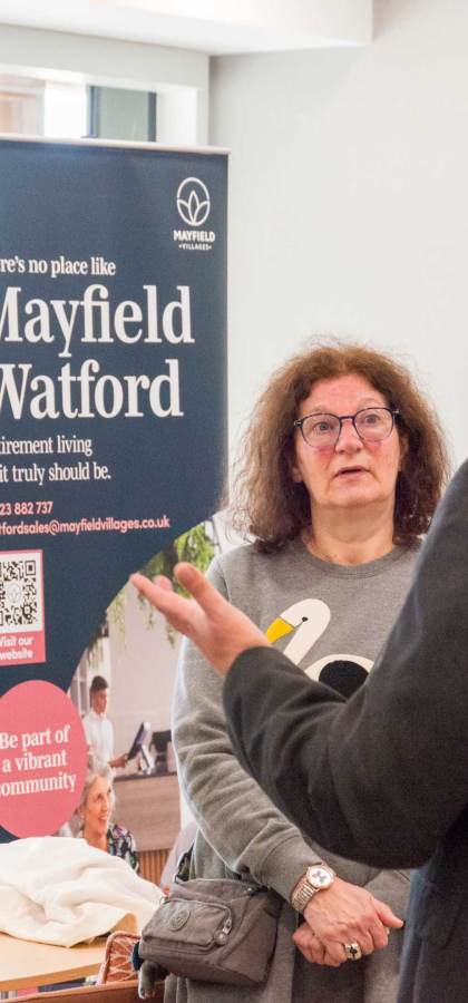 People standing indoors having a conversation near a banner for Mayfield Watford Retirement Living.