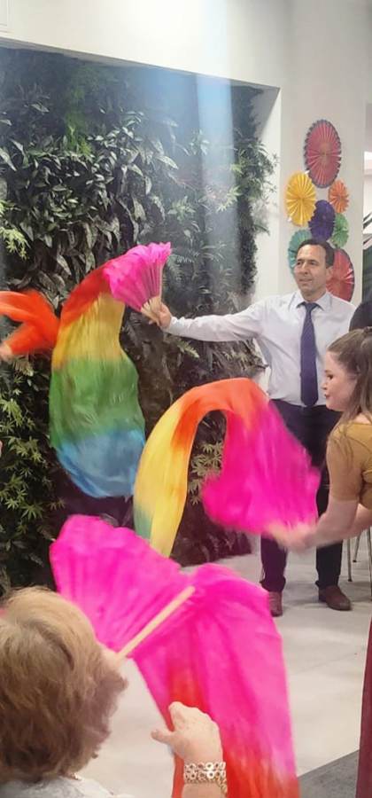 A group of people wave colorful rainbow scarves while others watch in a decorated indoor space with tables and greenery.