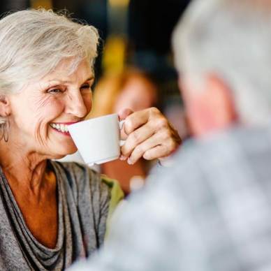 Elderly woman smiling and holding a cup, sitting across from a man.