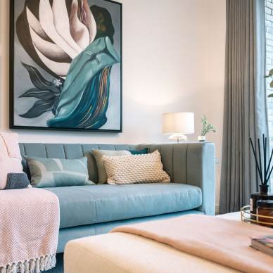 A modern living room featuring a teal sofa with cushions and a beige throw, a wall with abstract artwork, a lit table lamp, and a coffee table with decor items including a pink vase and reed diffuser.