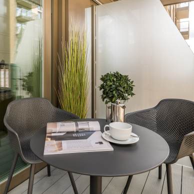 A small outdoor balcony with two black chairs and a round table. On the table are a white cup and saucer, two books, and a plant in a silver pot. The setup overlooks a building.