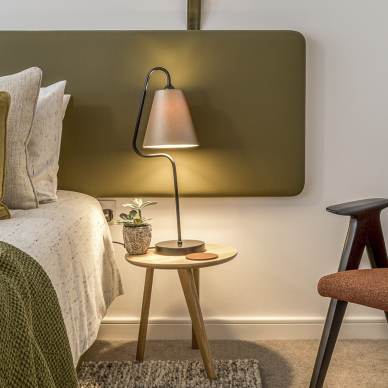A modern bedroom features a neatly made bed, a bedside table with a lamp and a small plant, and a brown upholstered chair. The room has a minimalist design with earthy tones.