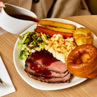 A person pours gravy onto a plate with roast beef, Yorkshire pudding, roast potatoes, carrots, cabbage, and cauliflower cheese. A fork and knife are on a napkin beside the plate.