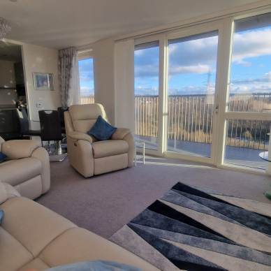 A bright living room with reclining chairs, a geometric rug, and large windows overlooking a scenic view. There's a dining area in the background and a side table with a lamp.