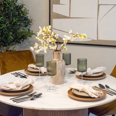 A round dining table set for four with rustic plates, green glasses, and napkins. A vase with white flowers is in the center. Greenery and abstract art decorate the background.