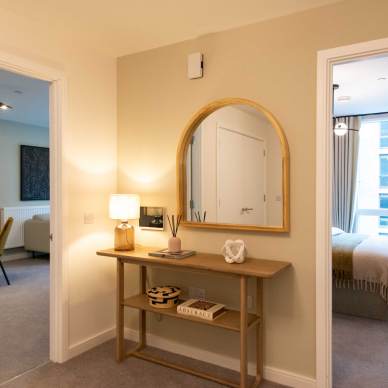 A hallway with a wooden console table and mirror. To the left is a dining area with a set table, and to the right is a bedroom with a made bed and large window.