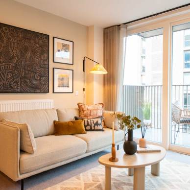 Modern living room with a beige sofa, patterned cushions, and a wooden coffee table. Art prints hang on the wall. A lamp illuminates the space. Balcony with seating is visible through glass doors.