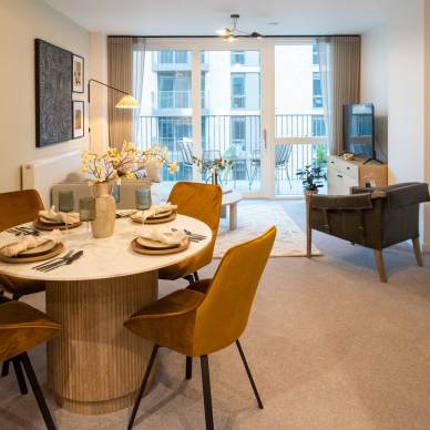 Modern open-plan living room and dining area with a round table, mustard chairs, and a TV. Large windows in the background offer a view of a balcony and buildings.