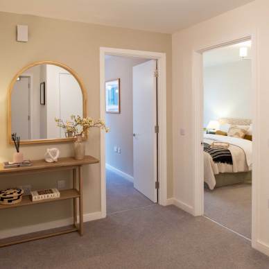 A hallway with a wooden console table, round mirror, and lamp. Two open doors lead to a bedroom and another room. Beige walls and carpeted floor.