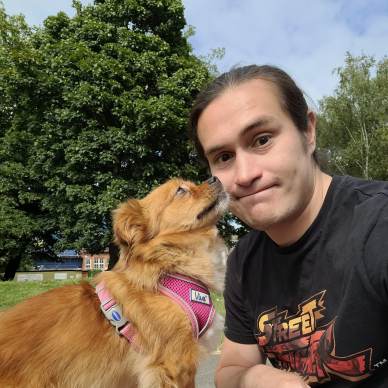 Person sitting outdoors with a small brown dog wearing a pink harness. Trees and sky in the background.