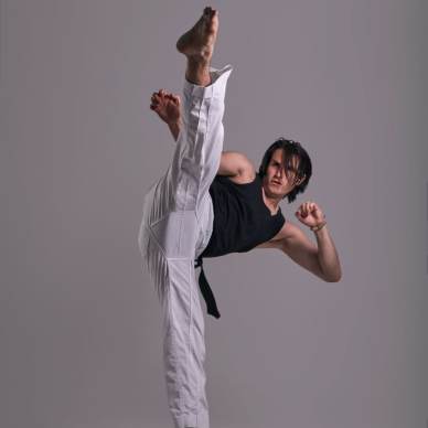 Person performing a high kick in martial arts attire, including a black belt and white pants, against a gray background.