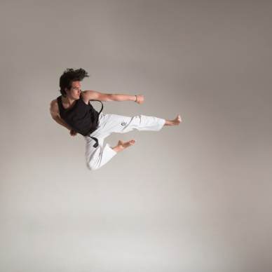 Person in a black shirt and white pants performing a mid-air martial arts kick against a plain background.