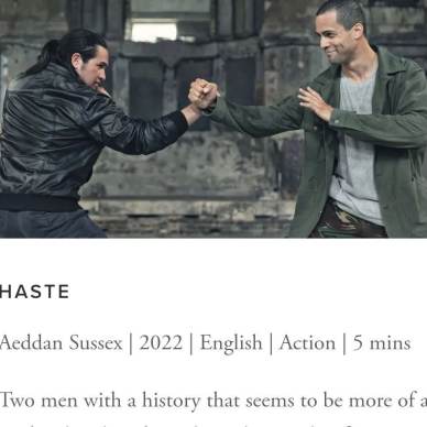 Two men in casual clothing engage in a choreographed fighting stance inside a rustic indoor setting.