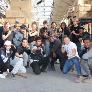 A group of people in casual clothing striking various poses in a warehouse with stacks of boxes in the background.