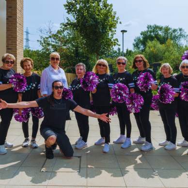 A group of women and one man poses outside, all dressed in black and holding purple pom-poms.