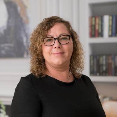 A woman with curly hair and glasses wearing a black top stands indoors in front of a bookcase and framed art.