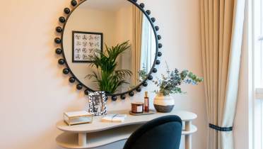 A stylish home office with a curved desk, black chair, and a round mirror reflecting a plant and wall art. A vase, candles, and books are on the desk. Beige curtains frame a window.