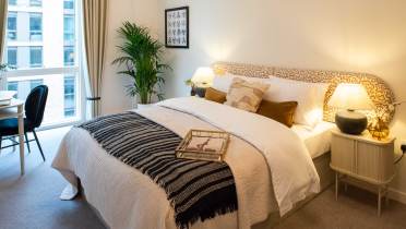 A neatly arranged bedroom with a double bed, patterned headboard, and pillows. A tray is on the bed. Plants, lamps, and a window with city view complete the decor.