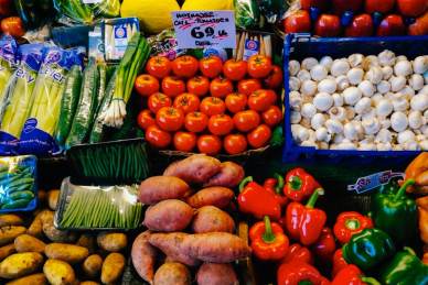 A variety of fresh vegetables are displayed for sale, including tomatoes, mushrooms, sweet potatoes, green onions, bell peppers, cucumbers, and potatoes.