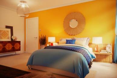 A cozy bedroom with a yellow accent wall, sunburst mirror, white door, wooden and blue bedding, bedside lamps, and a chest of drawers with various decor items.