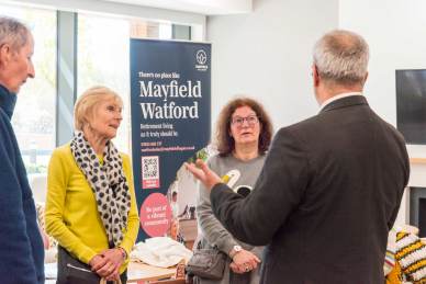 People standing indoors having a conversation near a banner for Mayfield Watford Retirement Living.