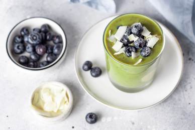 A glass of green smoothie topped with blueberries and coconut flakes on a white plate, with bowls of blueberries and coconut flakes nearby.