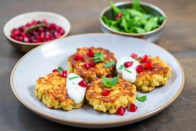 Three golden fritters topped with white sauce, pomegranate seeds, and parsley are served on a plate, with bowls of salad and pomegranate seeds in the background.