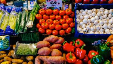 A variety of fresh vegetables are displayed for sale, including tomatoes, mushrooms, sweet potatoes, green onions, bell peppers, cucumbers, and potatoes.