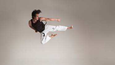 Person in a black shirt and white pants performing a mid-air martial arts kick against a plain background.