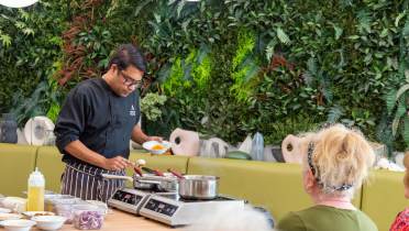 A chef demonstrates cooking at a table with ingredients, pans, and a hot plate, while people watch. The background features a lush, green wall.