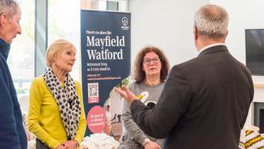 People standing indoors having a conversation near a banner for Mayfield Watford Retirement Living.