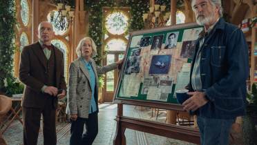 Three older adults stand beside a board covered with photos, documents, and maps in a sunlit room with plants and arched windows.