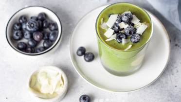 A glass of green smoothie topped with blueberries and coconut flakes on a white plate, with bowls of blueberries and coconut flakes nearby.